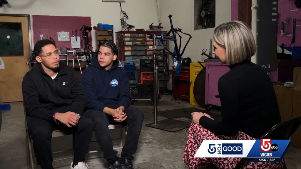 Two teenage boys seated facing an interviewer with bikes and work stations in the background.